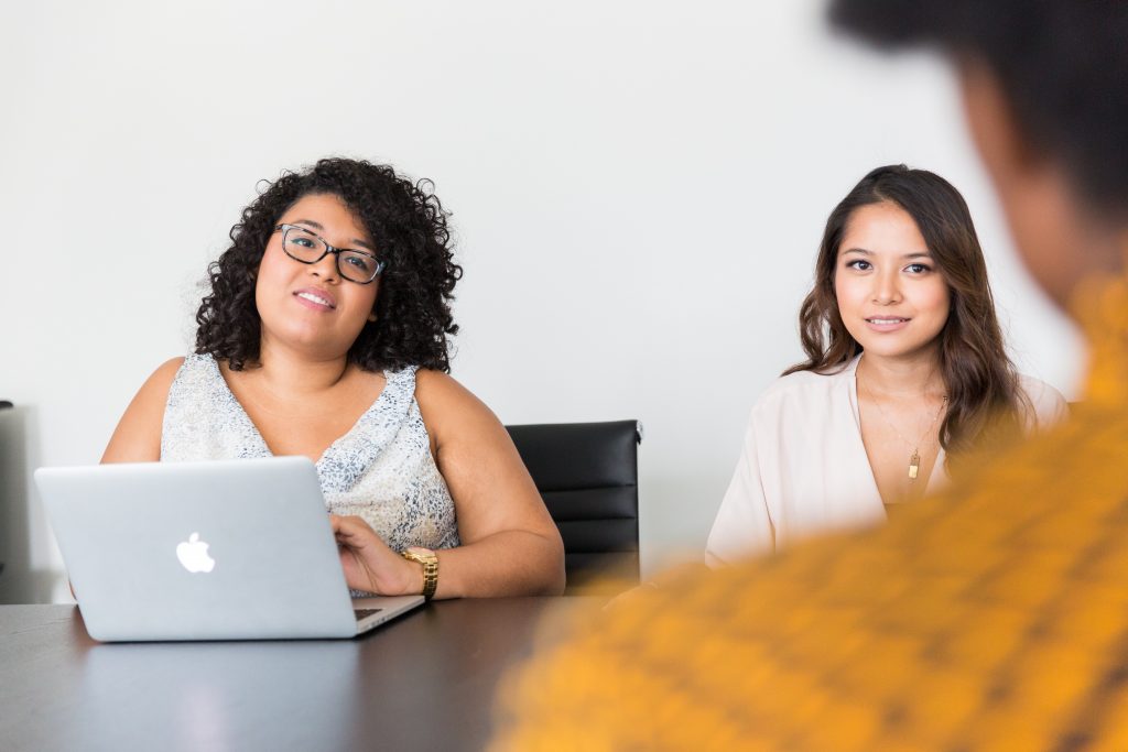 Image Description: Two racialized individuals staring a third person, while partially smiling. One of the two individuals has grey laptop in front of them.